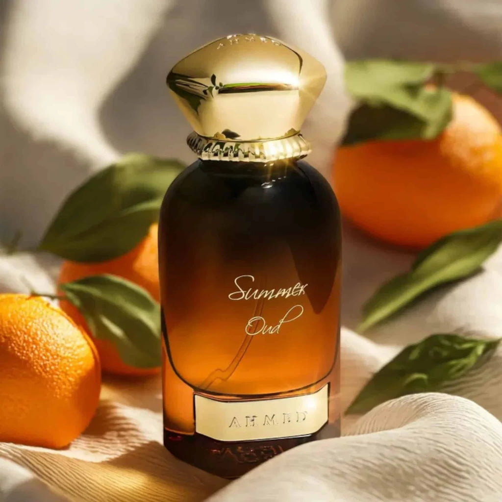 A bottle of A. Millia Summer Out perfume with a dark amber glass body and a shiny gold cap, styled with fresh oranges and green leaves on a soft beige fabric background.
