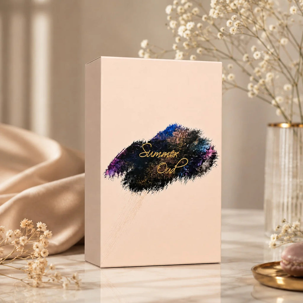A tall rectangular blush pink product box standing on a marble surface, decorated with a colorful lip-shaped graphic and the words 'Summer Out' in gold script. The box is styled in a warm indoor setting with beige satin fabric, dried flowers, and a glass vase in the background.